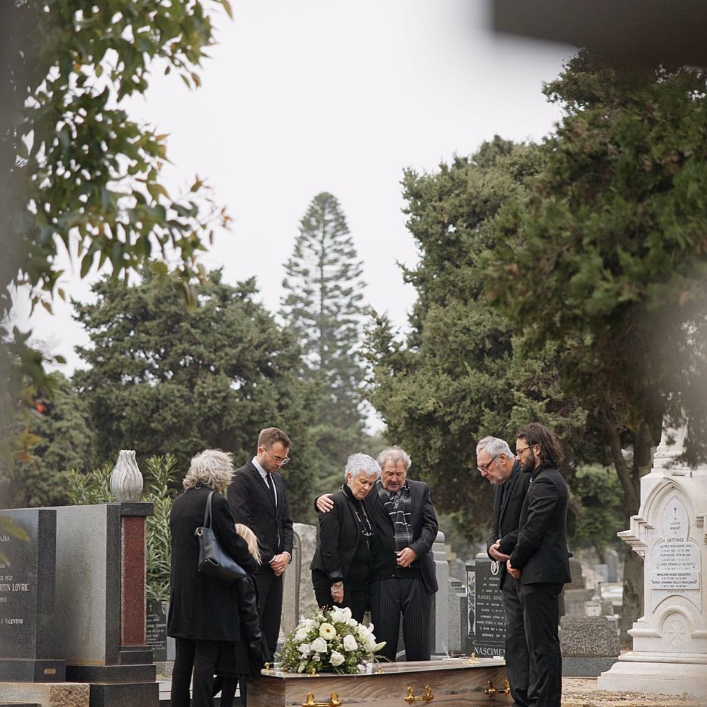 Dans un cimetière, un groupe de personnes vêtues de noir se tient autour d'un cercueil recouvert de fleurs blanches, la tête baissée en signe de deuil.