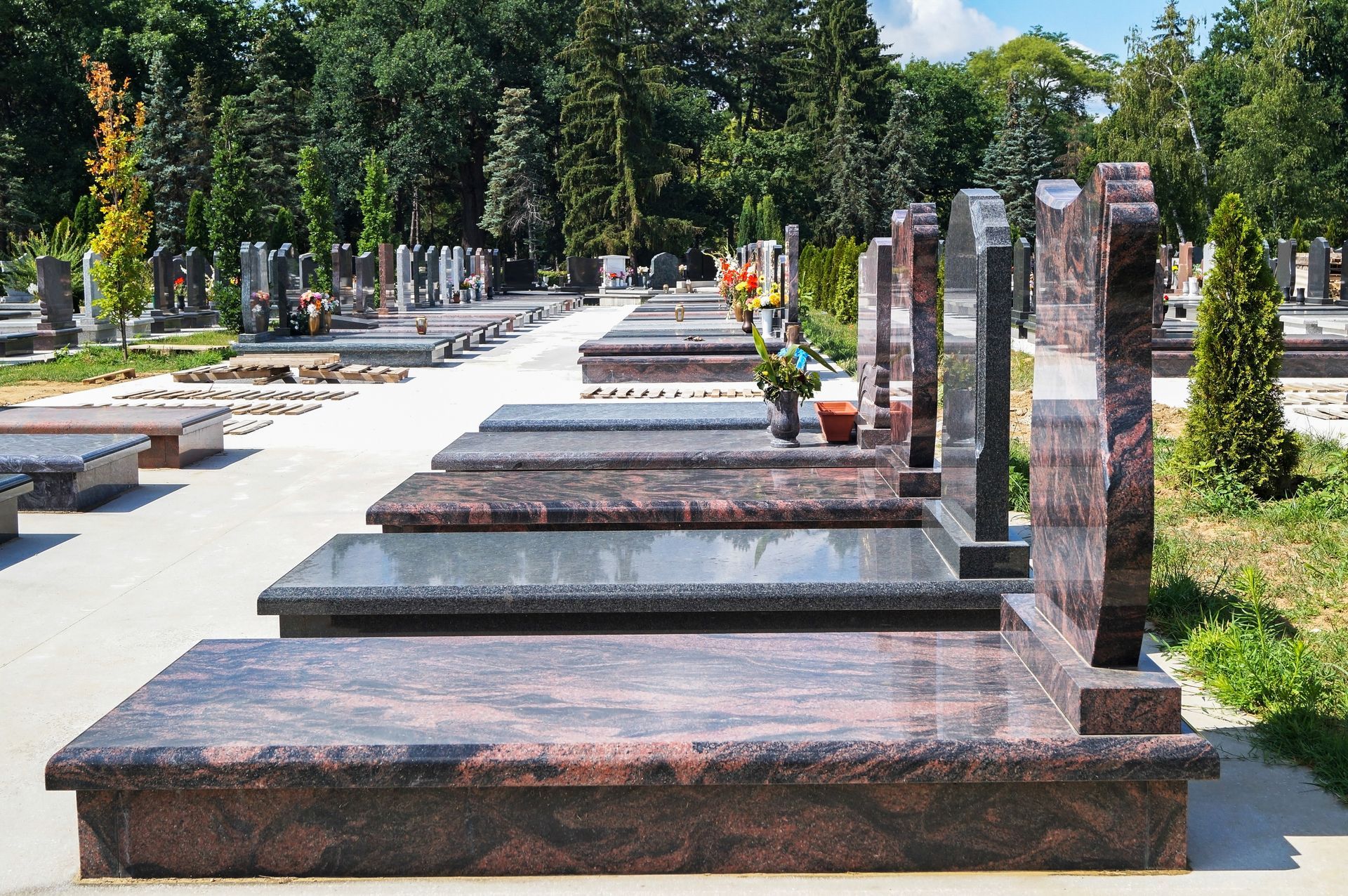 Des rangées de pierres tombales en granit poli dans un cimetière ensoleillé, arboré et bordé d'allées pavées.