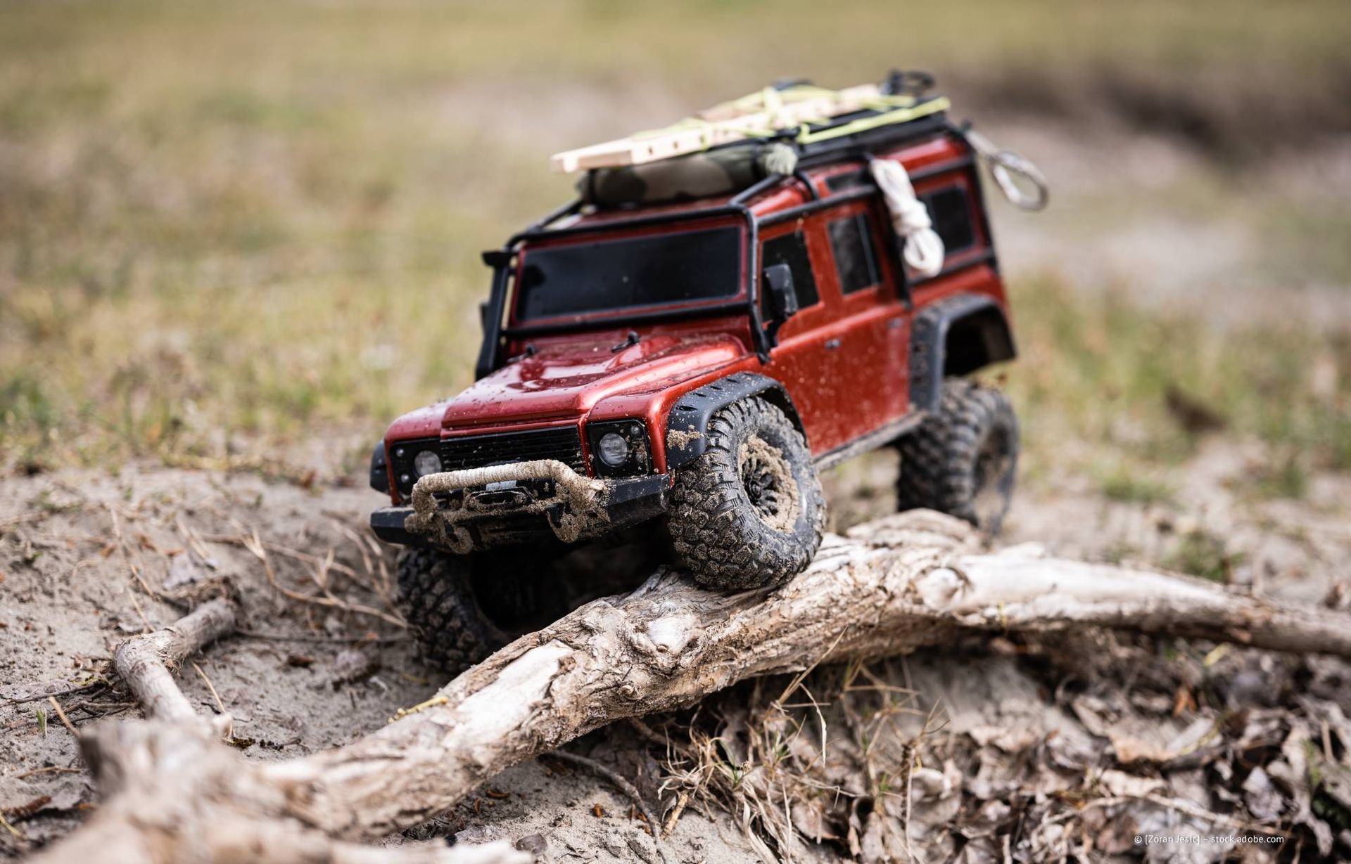 Ein roter Spielzeug-Land-Rover klettert auf einem Feldweg über einen Baumstamm.