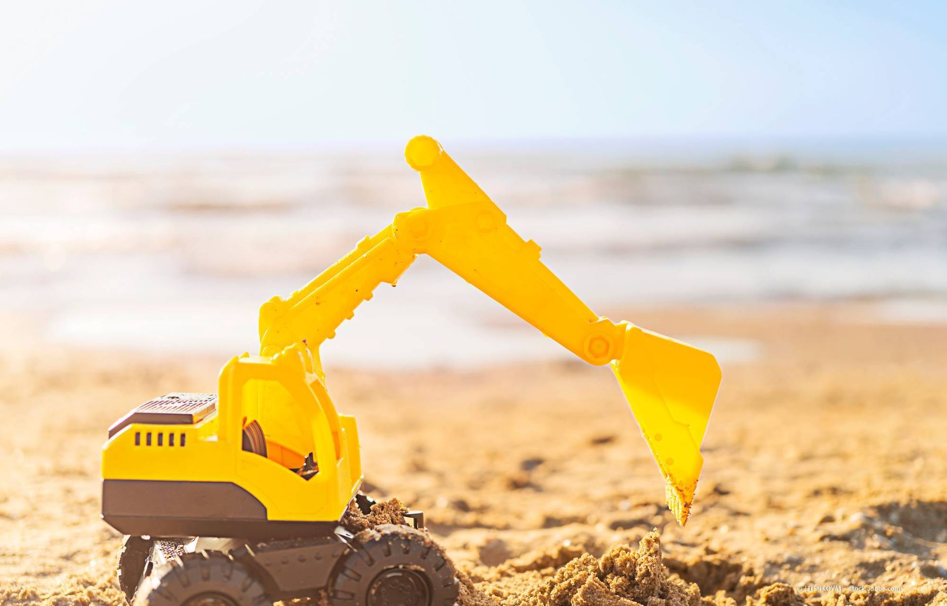 Gelber Spielzeugbagger im Sand am Strand, im Hintergrund das Meer.