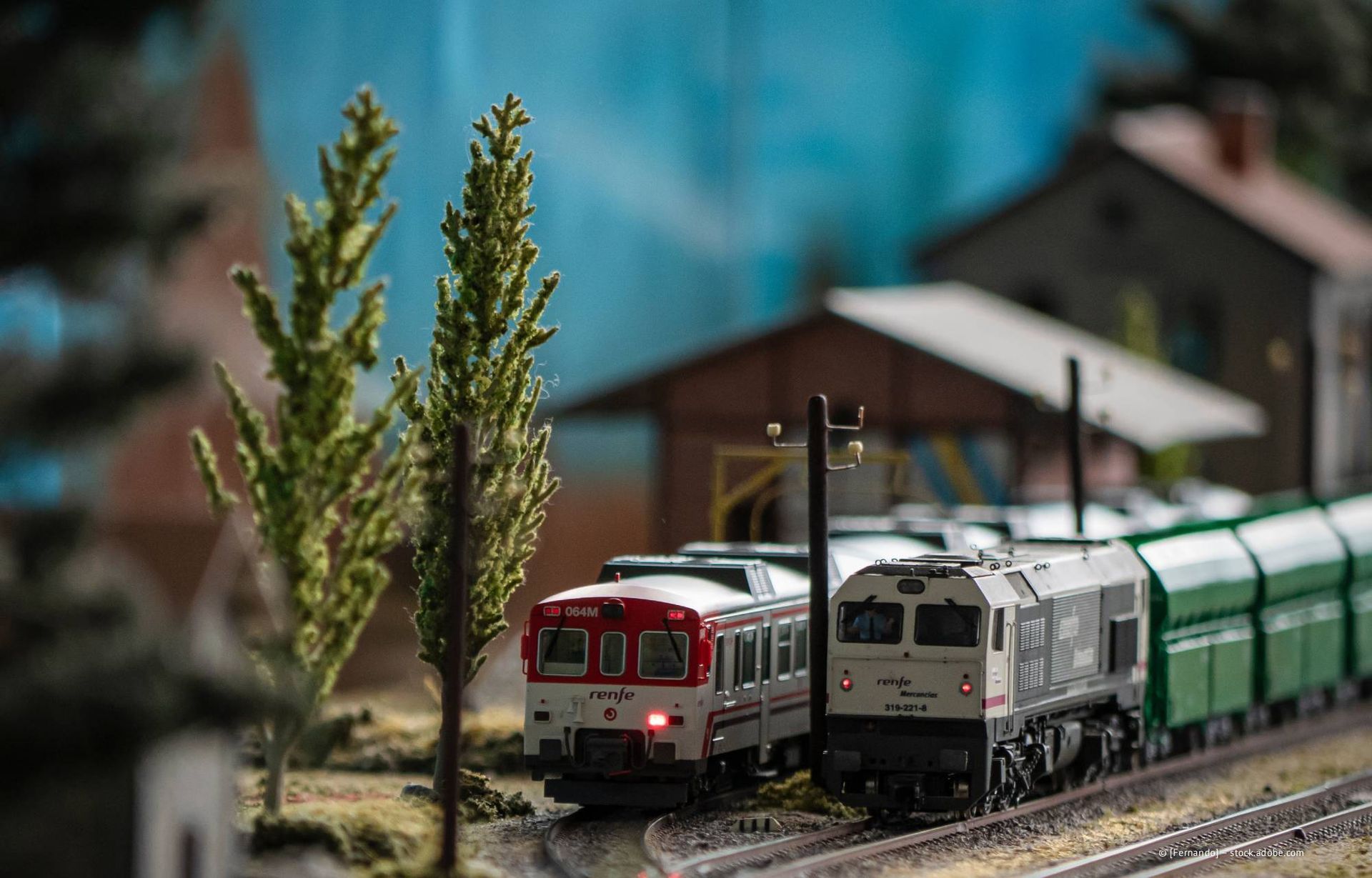 Modelleisenbahn mit rot-weißem Personenwagen und grauem Güterzug auf Gleisen, Bäume und Gebäude im Hintergrund.