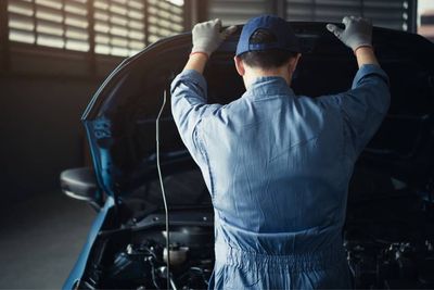 Un mécanicien en combinaison bleue travaille sur une voiture, capot ouvert, dans un garage faiblement éclairé.
