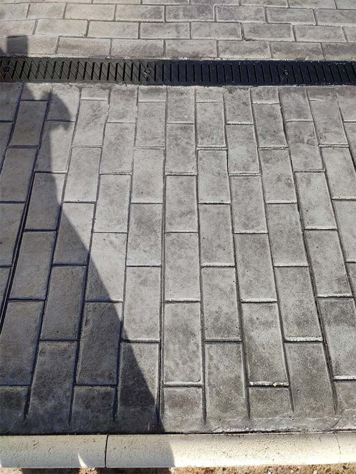 Pavimentos de ladrillo gris con desagüe lineal, zona sombreada y bordillo blanco.