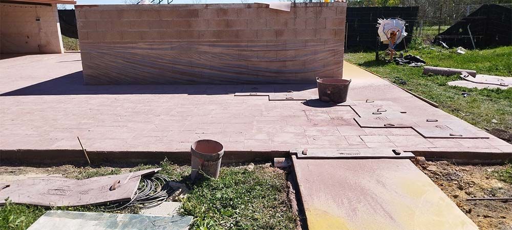 Obra con pavimento de ladrillo. Una gran estructura cuadrada se asienta sobre el pavimento, con varias baldosas. Se ven césped y un cubo.