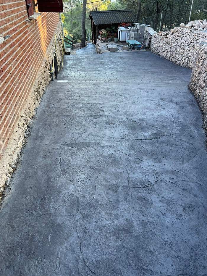 Camino de hormigón entre pared de ladrillo y pared de piedra, que conduce hacia una estructura y árboles.