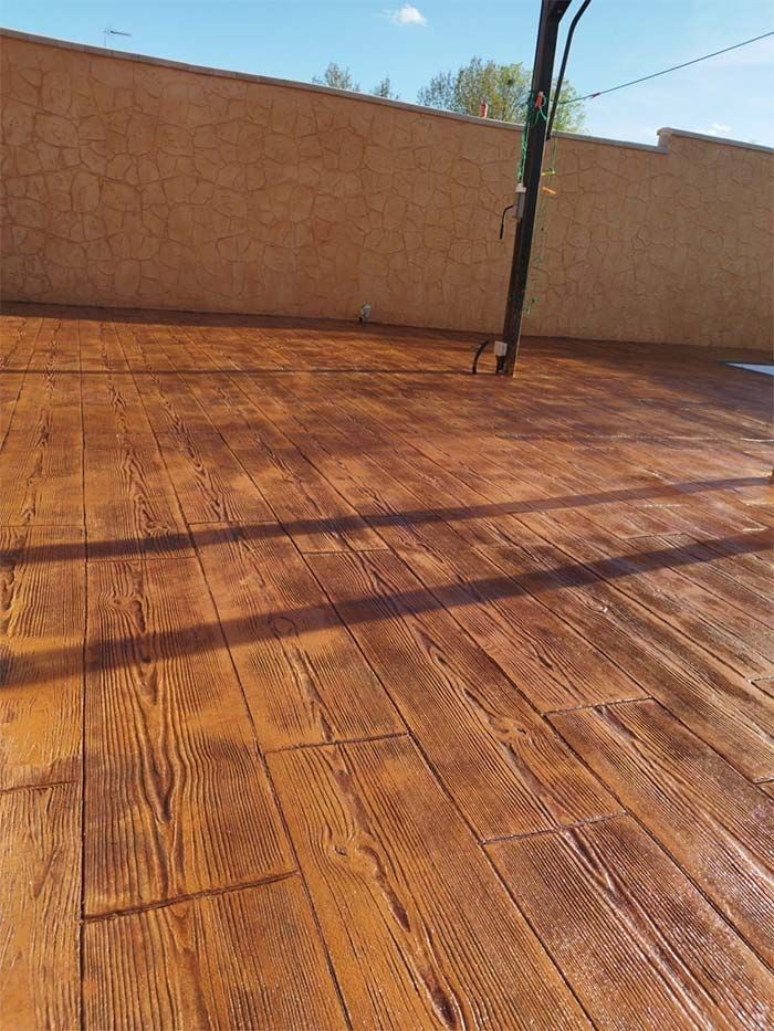 Patio de hormigón con diseño de tablones de madera con tinte marrón contra una pared texturizada de color beige bajo un cielo despejado.