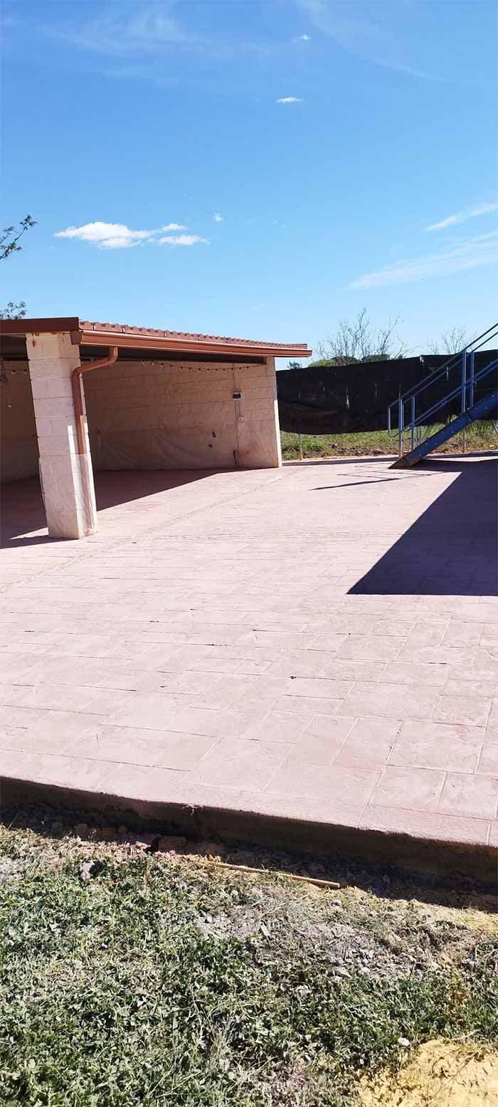 Un patio de hormigón con un pequeño refugio de ladrillos, rodeado de césped, bajo un cielo azul.