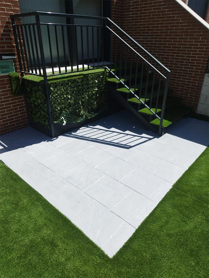 Patio exterior con baldosas grises, césped artificial verde, escaleras de metal negro y un muro de seto.