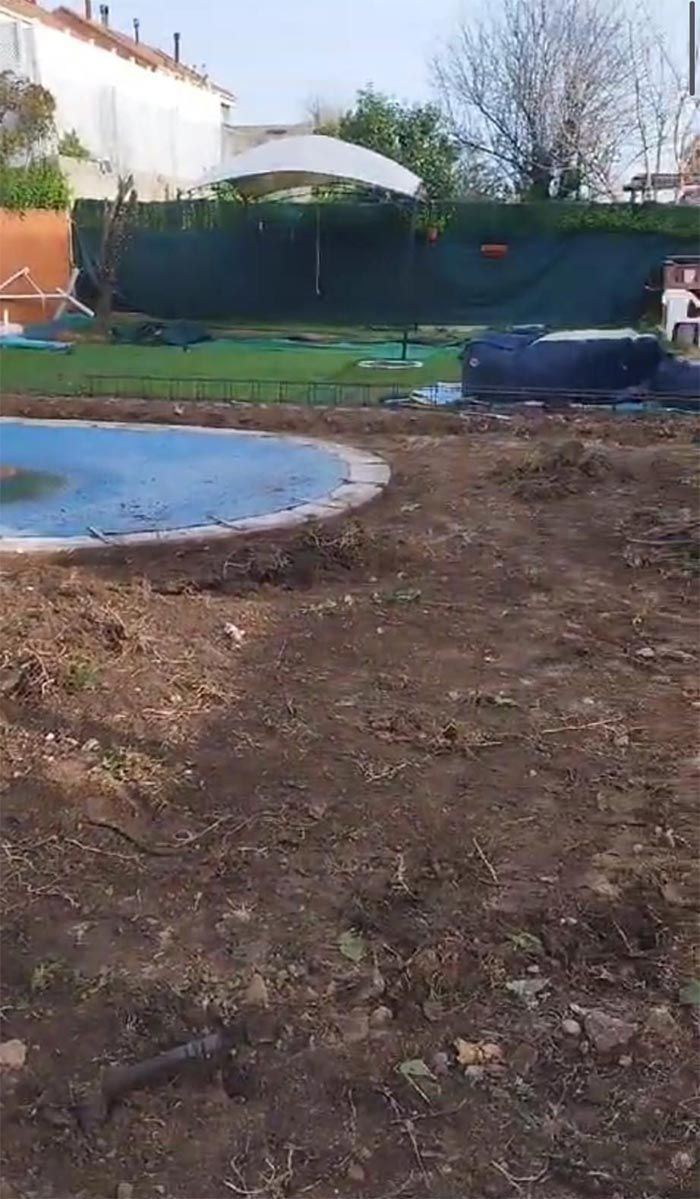 Zona de tierra con piscina curva. Césped verde y valla al fondo. Cielo nublado.