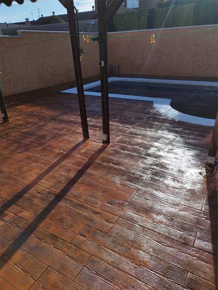 Patio de hormigón estampado marrón con pérgola, parcialmente húmedo, rodeando una piscina oscura.