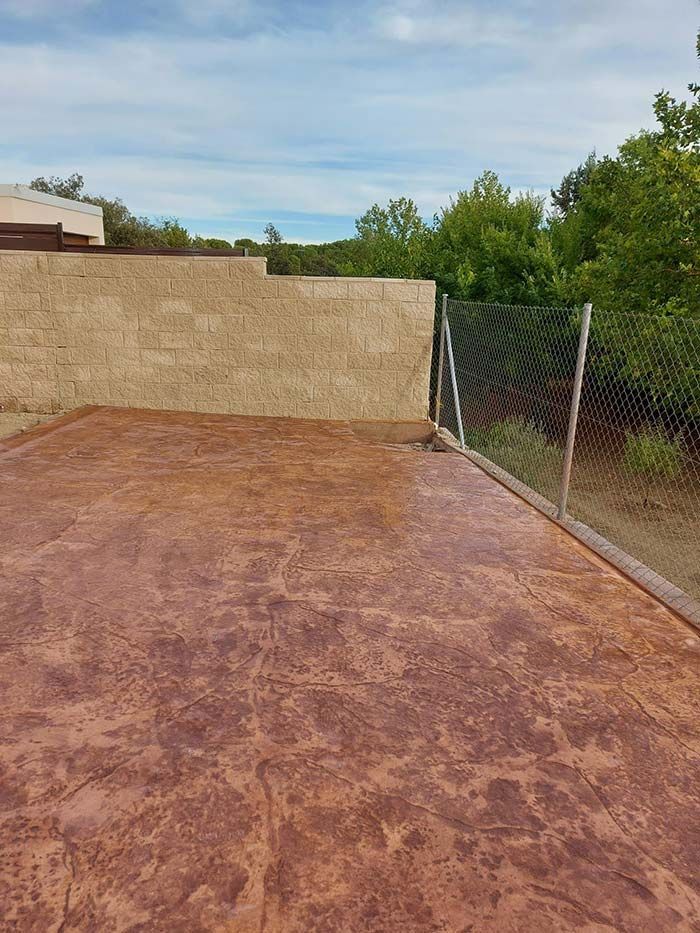 Patio de hormigón estampado de color marrón rojizo junto a una pared de estuco color tostado y una cerca de alambre, con árboles y un cielo azul de fondo.