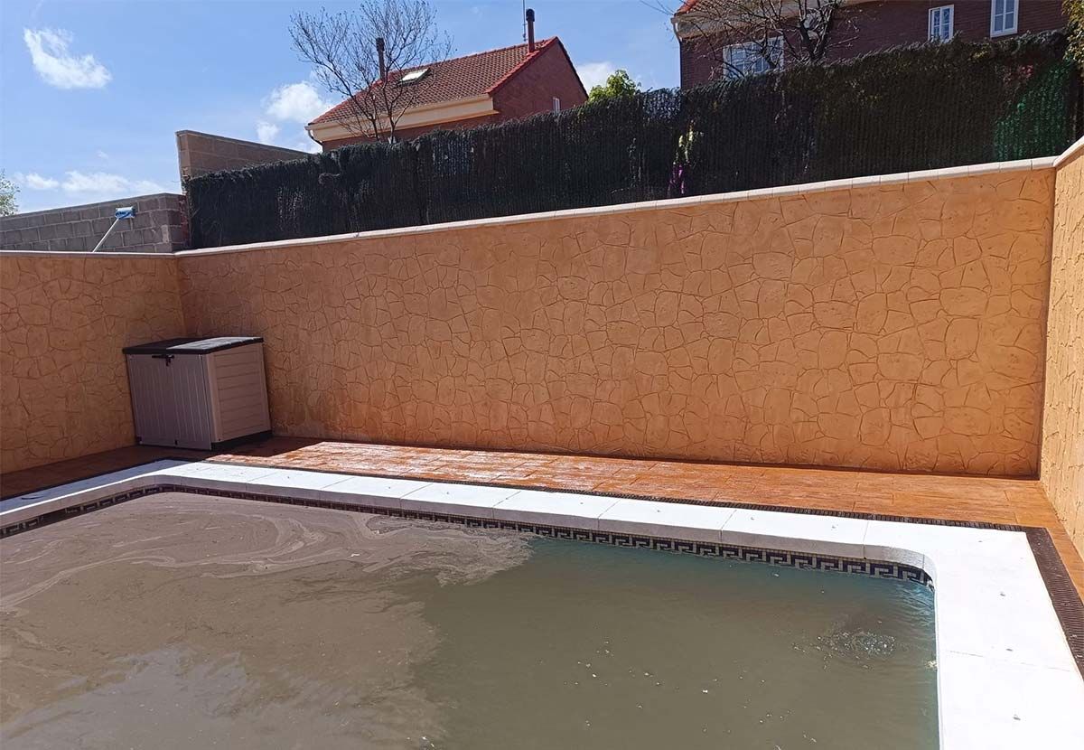 Una piscina en el patio trasero con paredes de estuco naranja y un seto, bajo un cielo brillante.