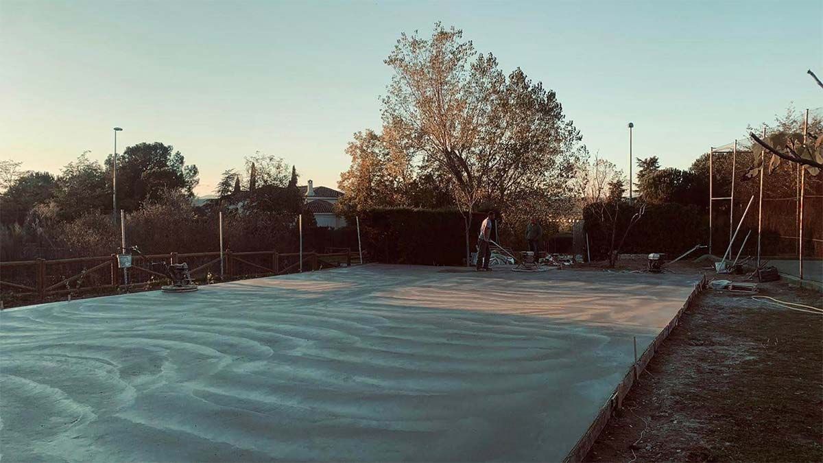 Superficie de hormigón recién vertido con vetas, rodeada de vallas, árboles y una persona en la distancia.