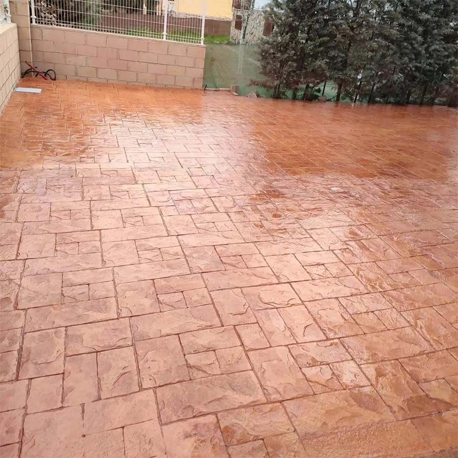 Patio húmedo de concreto estampado de color marrón rojizo con patrón de bloques, cerca de un muro de contención y árboles.