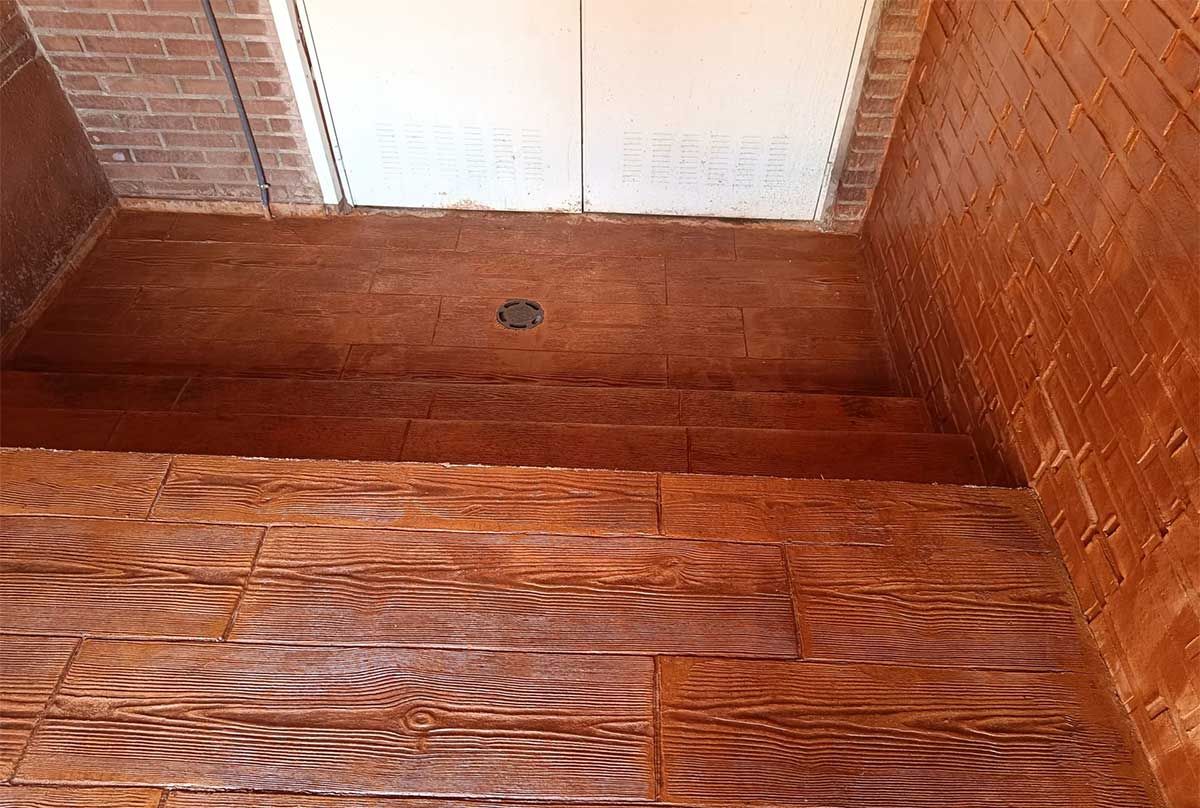 Suelo y paredes de imitación madera marrón con desagüe. La parte superior presenta un panel claro. Pared de ladrillo en la esquina superior izquierda.