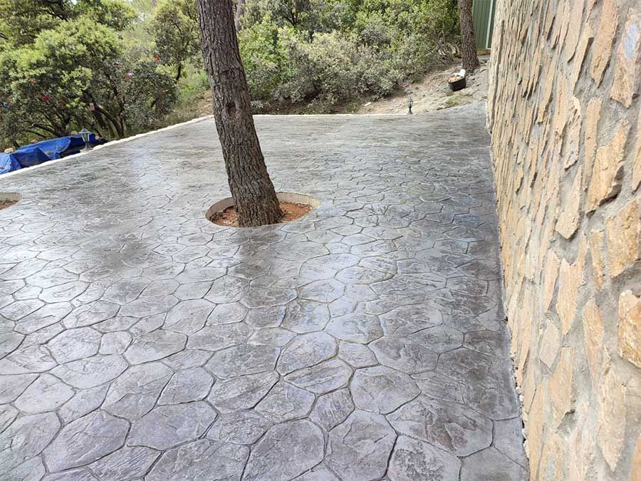 Patio de hormigón estampado con un árbol en el centro, junto a un muro de piedra, rodeado de arboles.