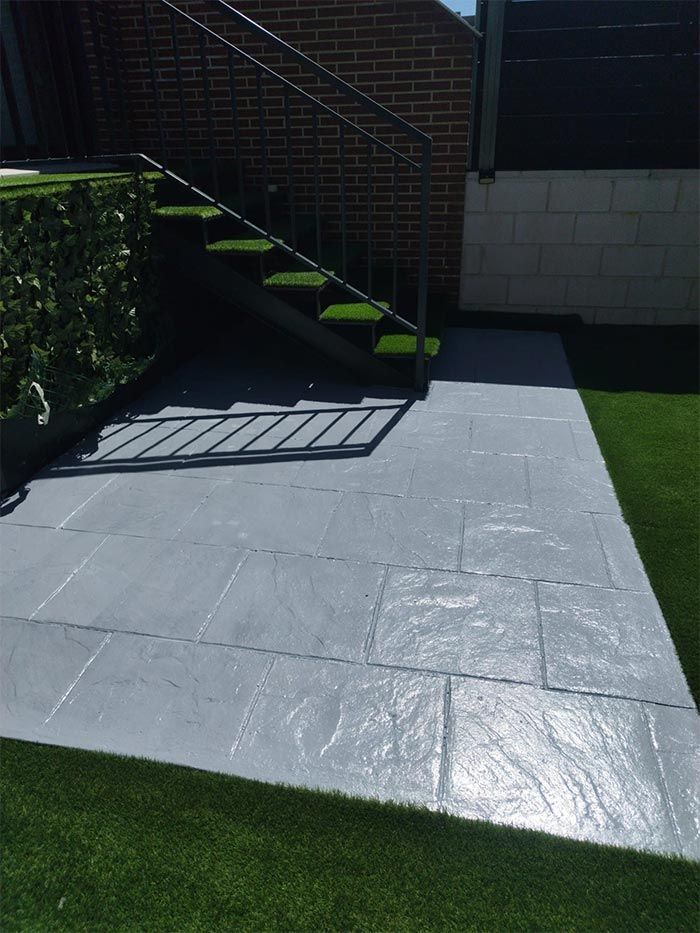 Patio pavimentado de color gris con escalones que suben, bordeado por césped verde artificial y una pared de ladrillos.