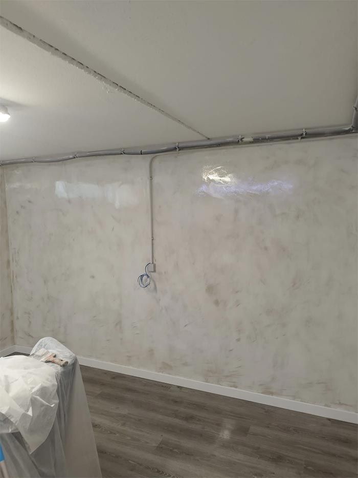 Una pared interior terminada con yeso brillante y texturizado en tonos blanquecino y marrón. Conducto eléctrico y zócalo gris.