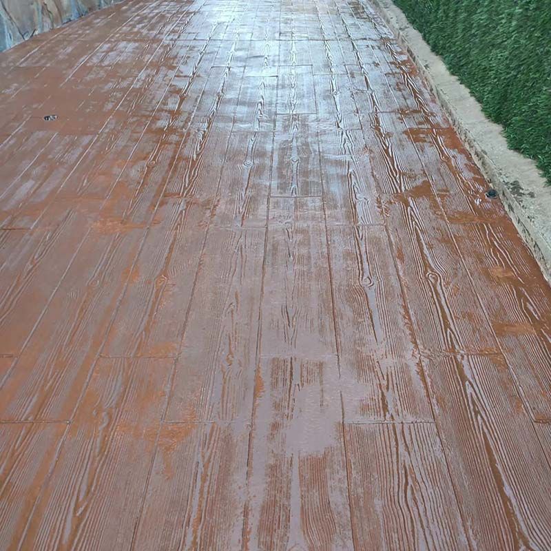 Camino de hormigón marrón con patrón de vetas de madera; un seto verde bordea el camino.