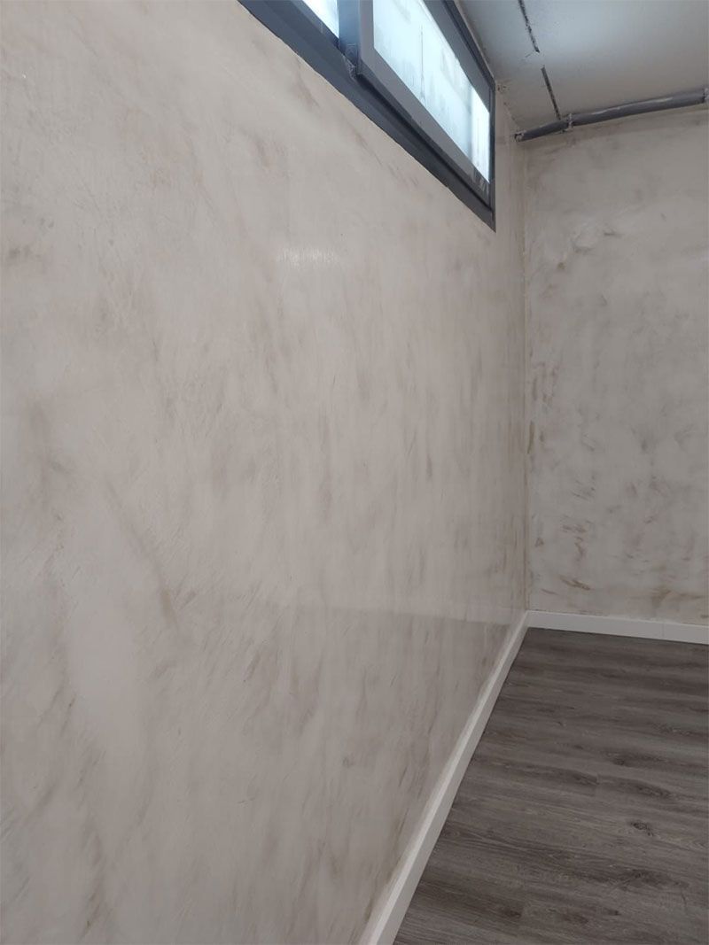 Habitación con paredes texturizadas de color claro, molduras grises y una ventana. Suelo de madera visible.