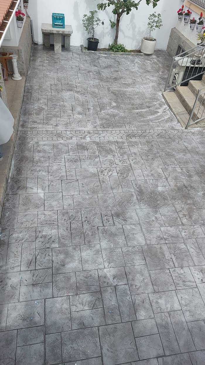 Patio de ladrillo gris, al aire libre. Se ven plantas, paredes blancas y un banco de piedra.