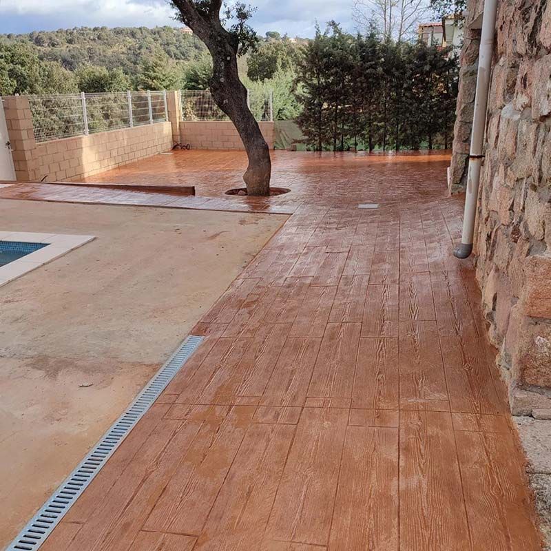 Espacio exterior con suelo de hormigón estampado en tonos marrones, arbol, piscina y pared de piedra.