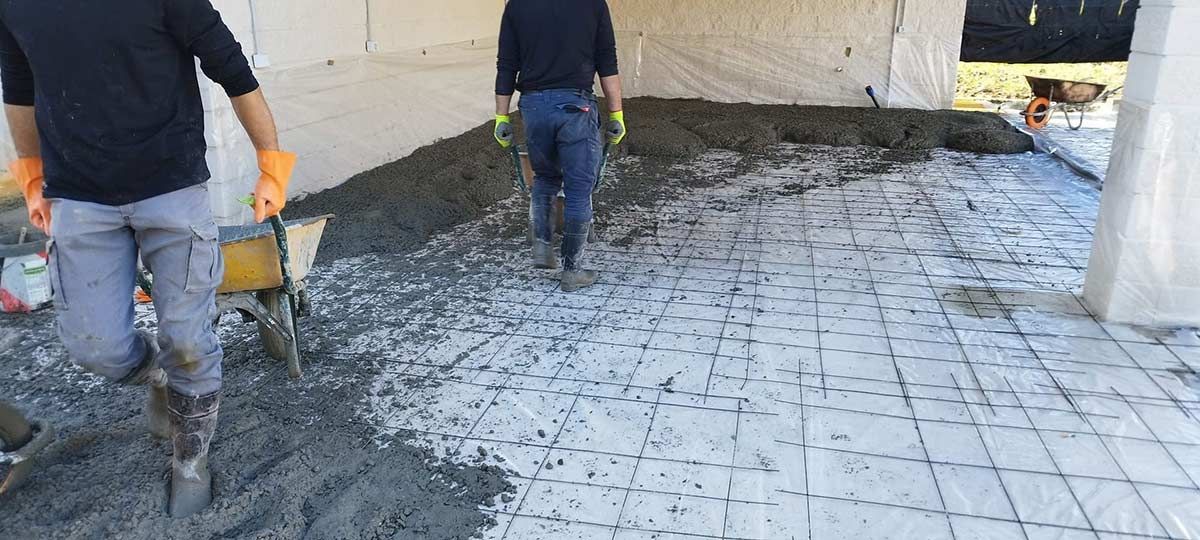 Obreros vierten hormigón sobre una superficie cuadriculada bajo una carpa. Llevan ropa de trabajo y guantes.