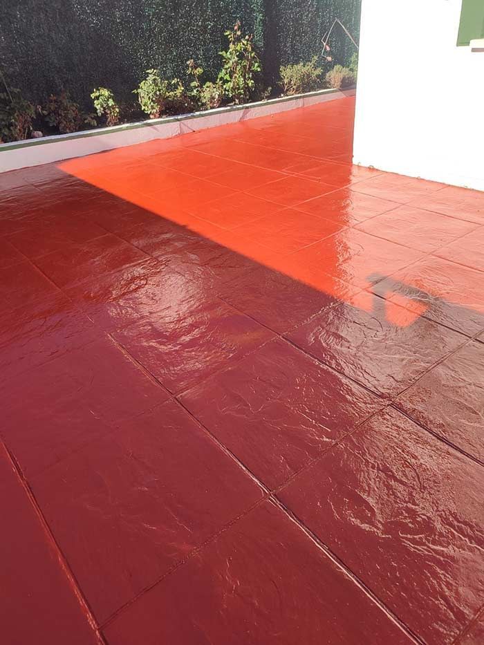 Deck pintado de rojo con zonas soleadas y sombreadas, adyacente a una pared blanca y arbustos verdes.