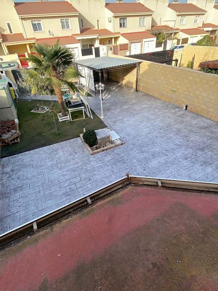 Vista aérea de un patio trasero suburbano con patio de concreto, césped, árboles pequeños y casas al fondo.