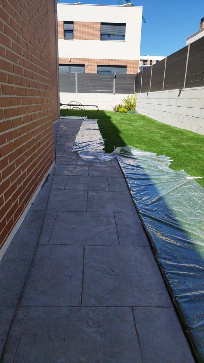 Un camino pavimentado a lo largo de una pared de ladrillos, que conduce hacia una zona de césped y una cerca en un día soleado.