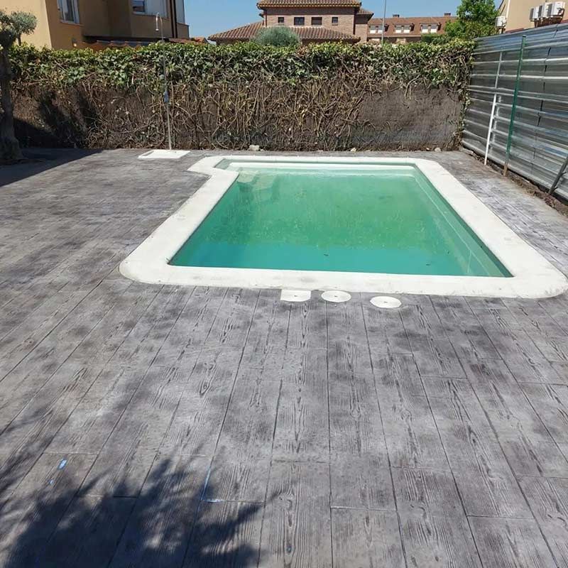 Piscina rectangular con borde blanco rodeada de hormigón gris, frente a un seto y una valla.