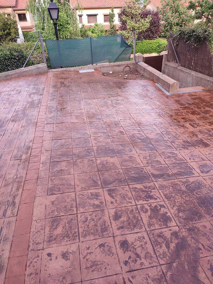 Camino de entrada de hormigón estampado con un patrón similar al ladrillo, que conduce a una cerca verde y edificios en el fondo.