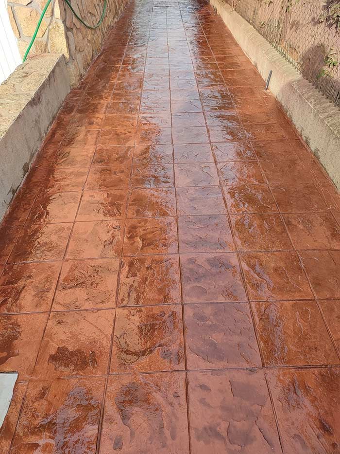 Pasarela de hormigón impreso, color marrón rojizo, con dibujo de cuadrados, entre dos muros bajos de hormigón.