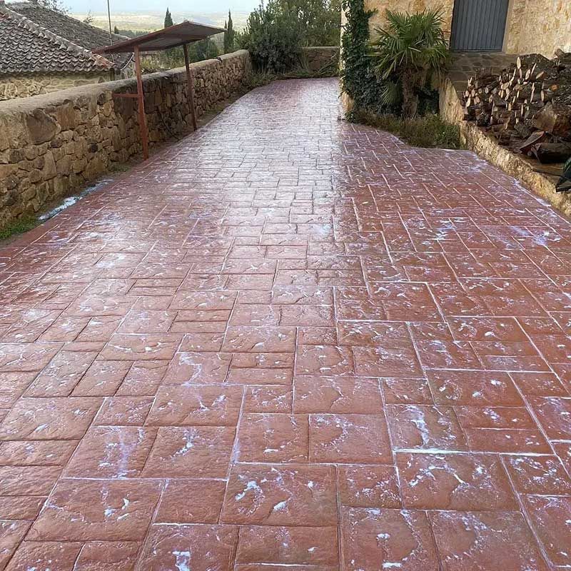 Camino pavimentado de color marrón rojizo con diseño de ladrillo y manchas de color claro, flanqueado por un muro de piedra y vegetación.