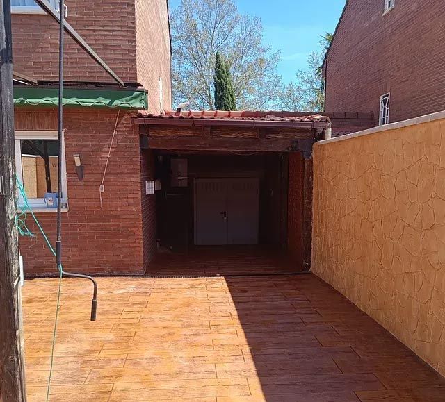 Garaje de ladrillo con puerta abierta, camino de entrada de hormigón estampado marrón, flanqueado por paredes de ladrillo y estuco, bajo un cielo azul.