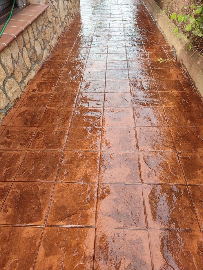 Pasarela de hormigón estampado, húmedo, de color marrón rojizo, con patrón de baldosas, bordeada por un muro de piedra y vegetación.