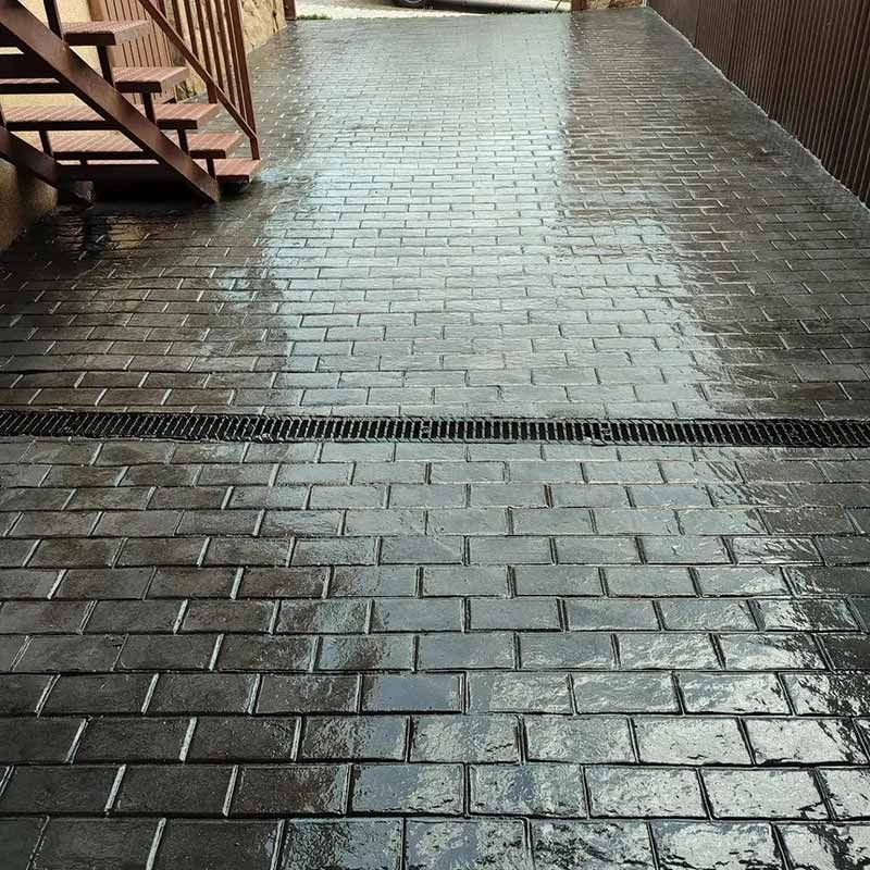 Camino de entrada de ladrillo oscuro y húmedo con desagüe, cerca de escaleras de madera y una cerca.