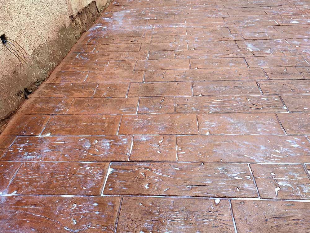 Patio de hormigón de color marrón rojizo con textura tipo ladrillo y eflorescencia blanca cerca de una pared de color marrón claro.