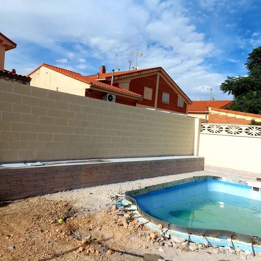 Una piscina en el patio trasero con una pared de ladrillos de color marrón y casas al fondo en un día parcialmente nublado.