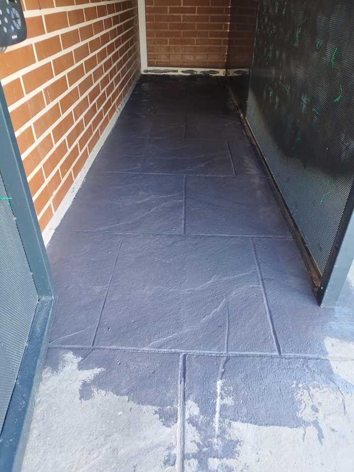 Piso de patio similar a piedra gris oscuro en un espacio exterior estrecho con una pared de ladrillo a la izquierda.