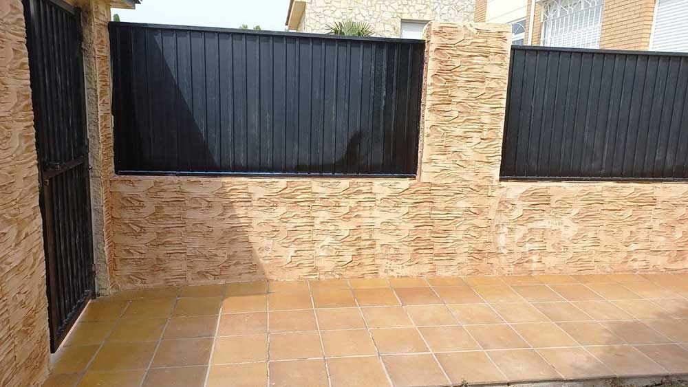 Sección de puerta y cerca de metal negro con pared texturizada en color bronce y patio embaldosado.