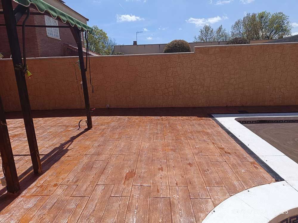 Patio trasero con hormigón estampado con apariencia de madera, una piscina y una estructura elevada contra una pared de estuco.