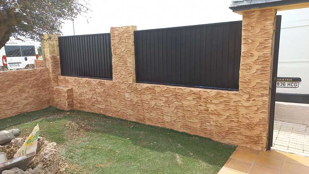 Valla de piedra y metal negro que bordea el césped. Furgoneta blanca al fondo.