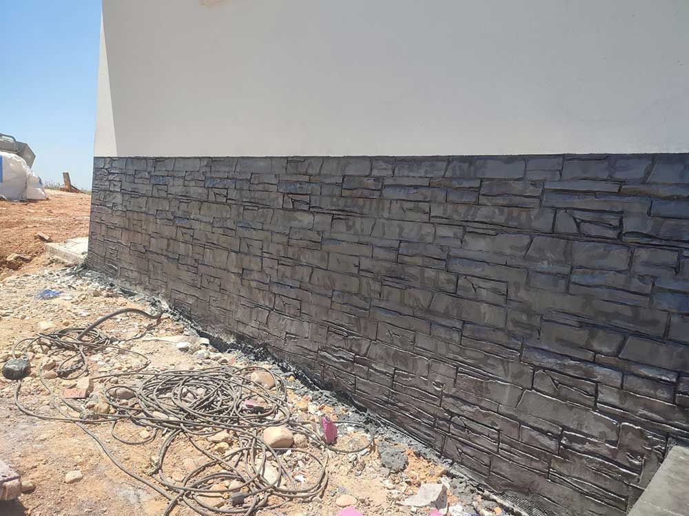 Base de pared de hormigón con textura de piedra de un edificio en construcción, bloques de color gris oscuro contra una pared blanca y suelo de tierra.