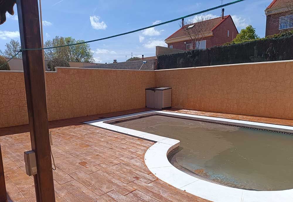 Un patio trasero con piscina, terraza marrón y una pared de estuco naranja en un día soleado.