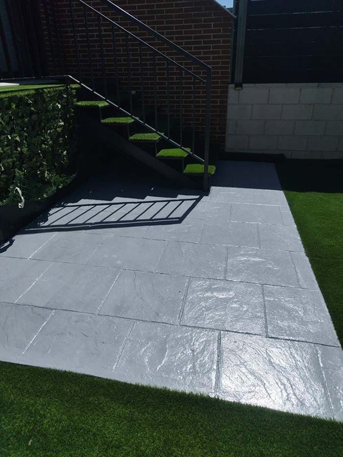 Patio gris con escaleras que suben, césped artificial verde y una pared de ladrillo.