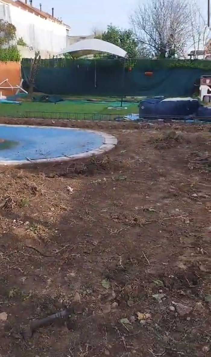 Parcela de tierra frente a una piscina de azulejos azules en construcción, respaldada por una valla verde y edificios.