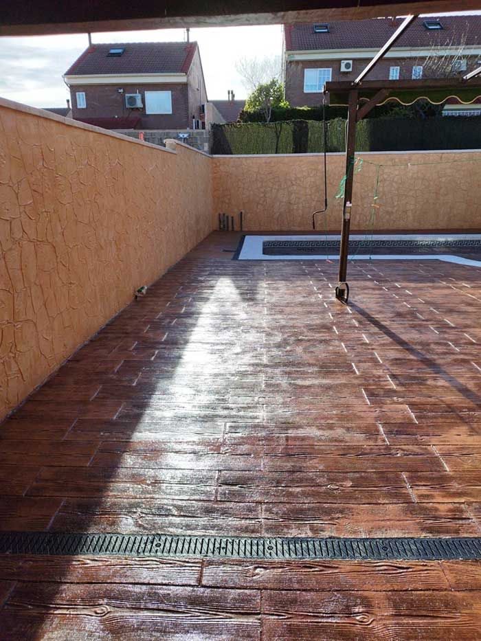 Patio de concreto estampado marrón con canal de drenaje, muro alto y casas de ladrillo de dos pisos al fondo.