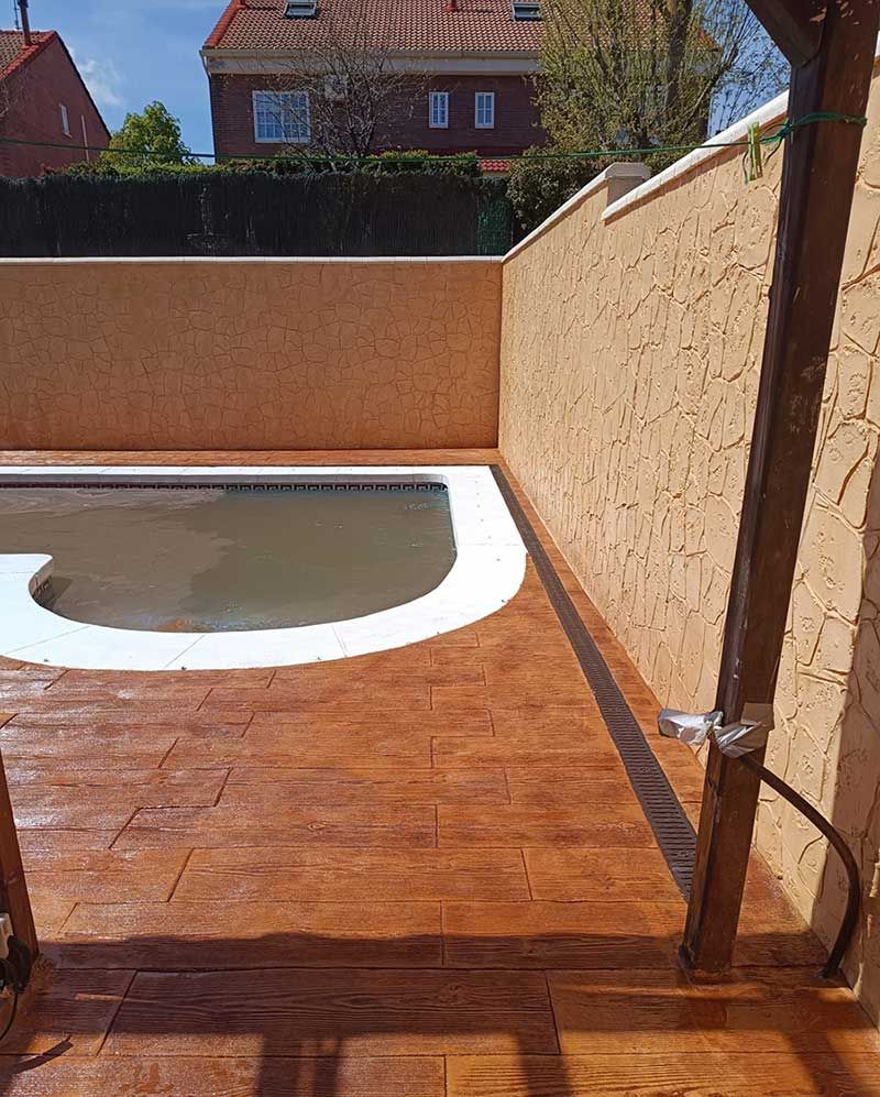 Área de piscina con piso de baldosas marrones, paredes de estuco color tostado y una piscina con borde blanco.