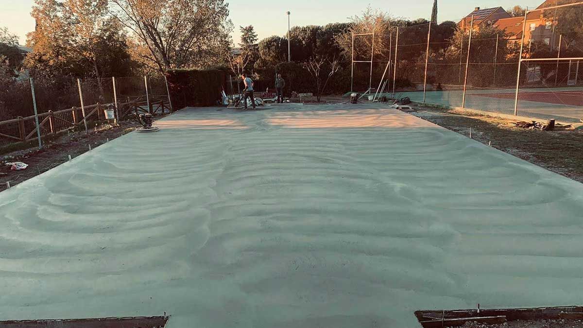 Superficie de hormigón recién vertido; trabajadores al fondo. Exterior con árboles y postes.